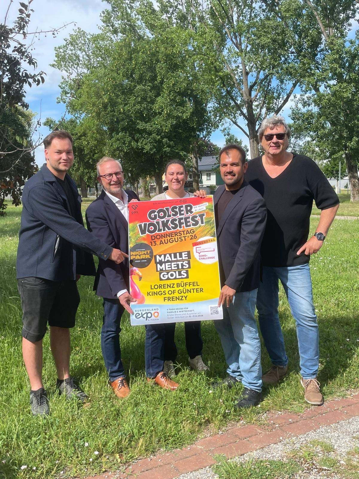 Fünf Personen stehen auf einem Grasfeld und halten ein großes Plakat. Das Plakat zeigt die Worte 'Goiserer Volksfest' und ein farbenfrohes Bild. Die Männer tragen formelle Kleidung, und einer trägt eine Sonnenbrille. Bäume und ein Gebäude sind im Hintergrund.