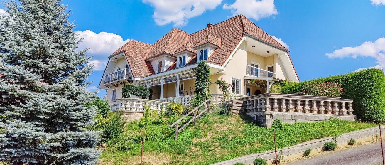 Ein großes, gut gepflegtes Haus mit einem braunen Dach und weißen Wänden, auf einem Hügel gelegen mit Balkon und Geländern. Das Haus ist von Grünpflanzen und einem hohen Tannenbaum umgeben.