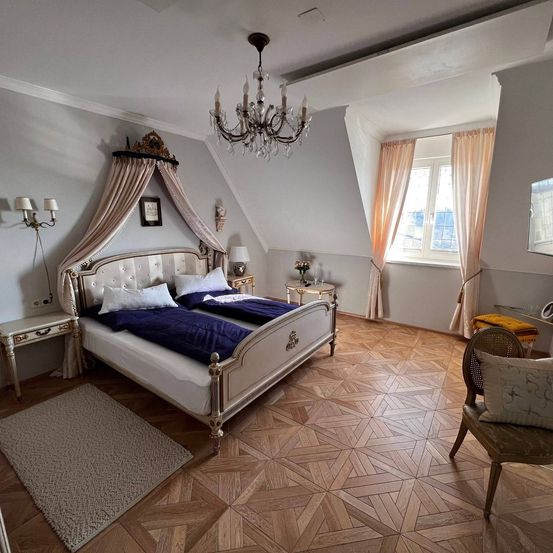 Ein Schlafzimmer mit einem großen Bett, einer Kristallleuchte und Holzparkettboden. Es gibt zwei Nachttische, einen Teppich und einen Stuhl. Das Zimmer hat ein Fenster mit Vorhängen.