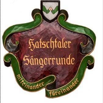 Ein roter Schild mit goldener Schrift in einem grünen Rahmen sagt 'Hatschaler Sangerrunde'. Darunter befindet sich ein grünes Banner mit den Worten 'Miteinander Fureinander'.