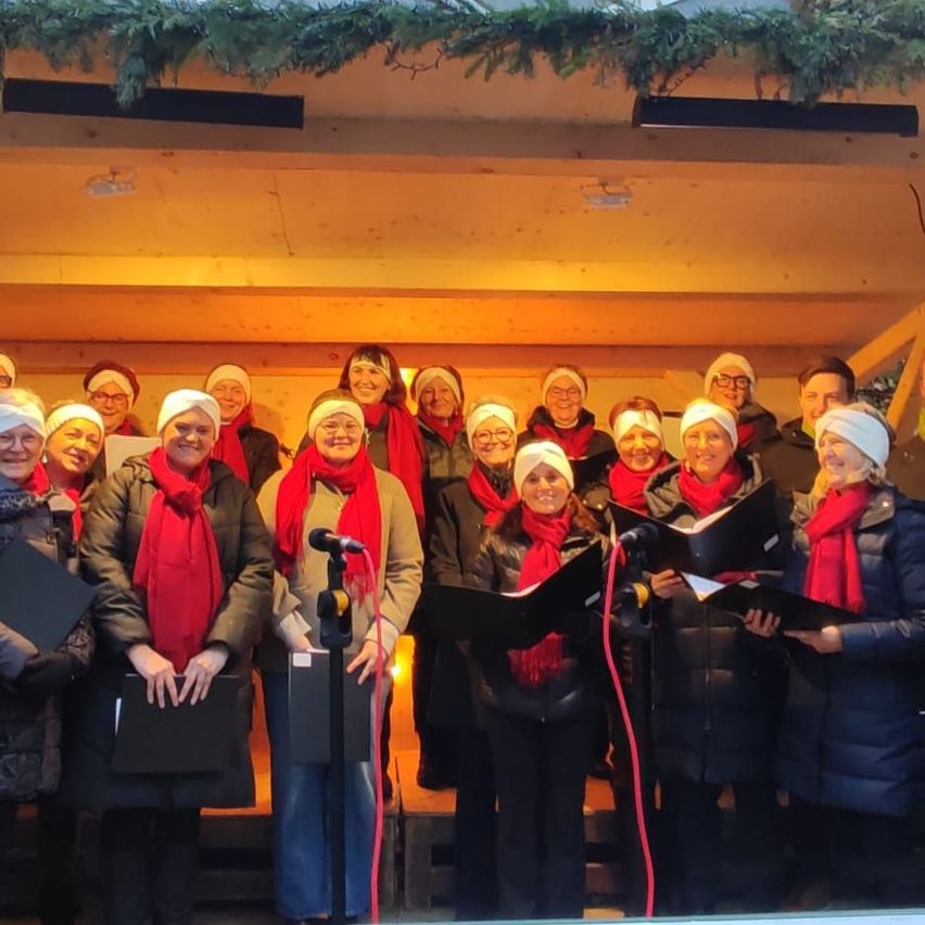 Ein Chor in Winterkleidung steht zusammen, hält Notenbücher und singt. Sie befinden sich in einem Raum mit Holzwänden und geschmückt mit Grün.