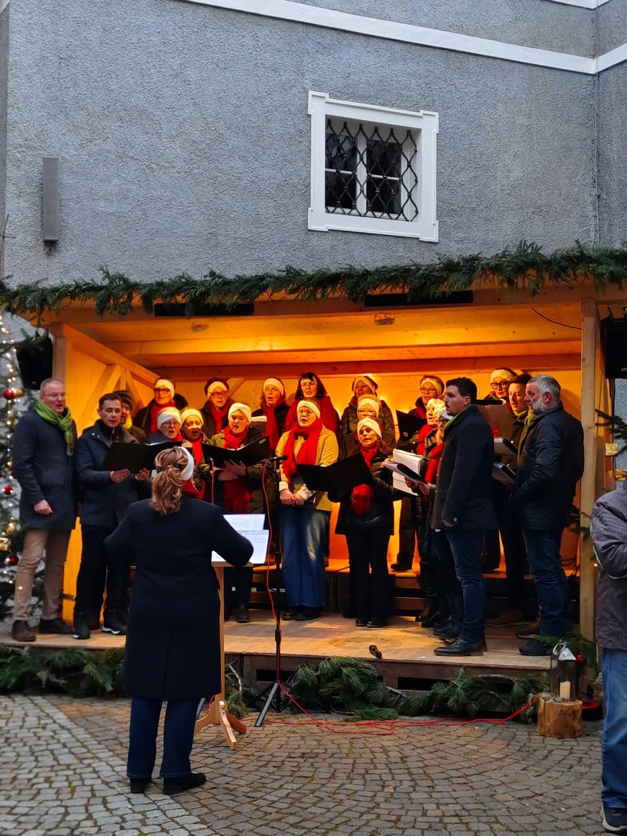 Ein Chor in festlicher Kleidung singt vor einem mit Weihnachtsdekoration geschmückten Gebäude. Der Chor steht auf einer hölzernen Bühne, hält Notenblätter und singt harmonisch.