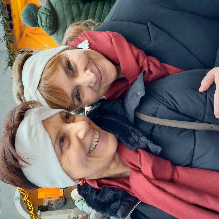 Zwei lächelnde Frauen mit Stirnbändern posieren eng für ein Selfie, wobei eine Frau ein rotes und die andere ein blaues Jackett trägt. Dahinter steht eine weitere Person in einem grünen Jackett und einem festlichen Hintergrund.