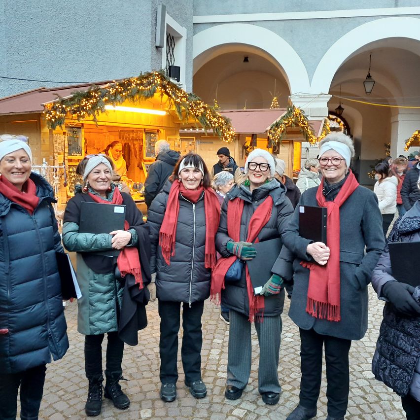 Fünf Frauen stehen vor einem geschmückten Laden und lächeln für ein Foto. Dahinter ist ein Laden mit Weihnachtslichtern und einem Weihnachtsbaum geschmückt. Andere Leute gehen im Hintergrund herum.