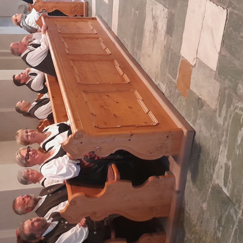 Eine Gruppe Erwachsener in traditioneller Kleidung sitzt auf hölzernen Kirchenbänken. Sie schauen zum Altar, und einige lächeln.