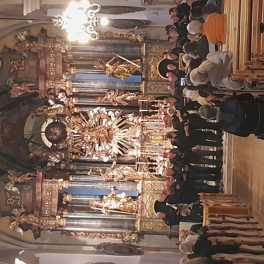 Eine Gruppe von Menschen versammelt sich in einer Kirche. Sie sehen einem wunderschön geschmückten Altar mit goldenen Statuen und kunstvollen Verzierungen gegenüber. Oben hängt eine Kronleuchter.