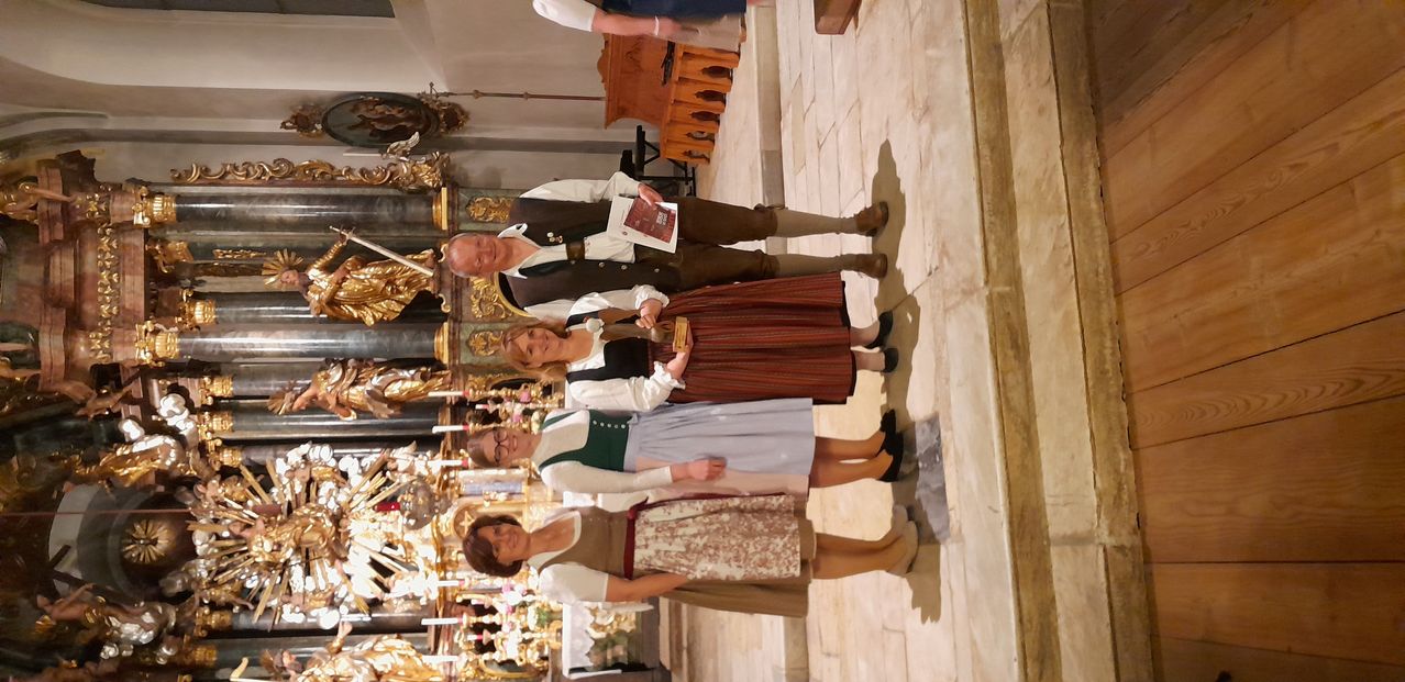 Fünf Menschen in traditioneller Kleidung stehen in einer Kirche. Drei Frauen und zwei Männer, einer hält ein Buch. Hinter ihnen ist ein Altar.