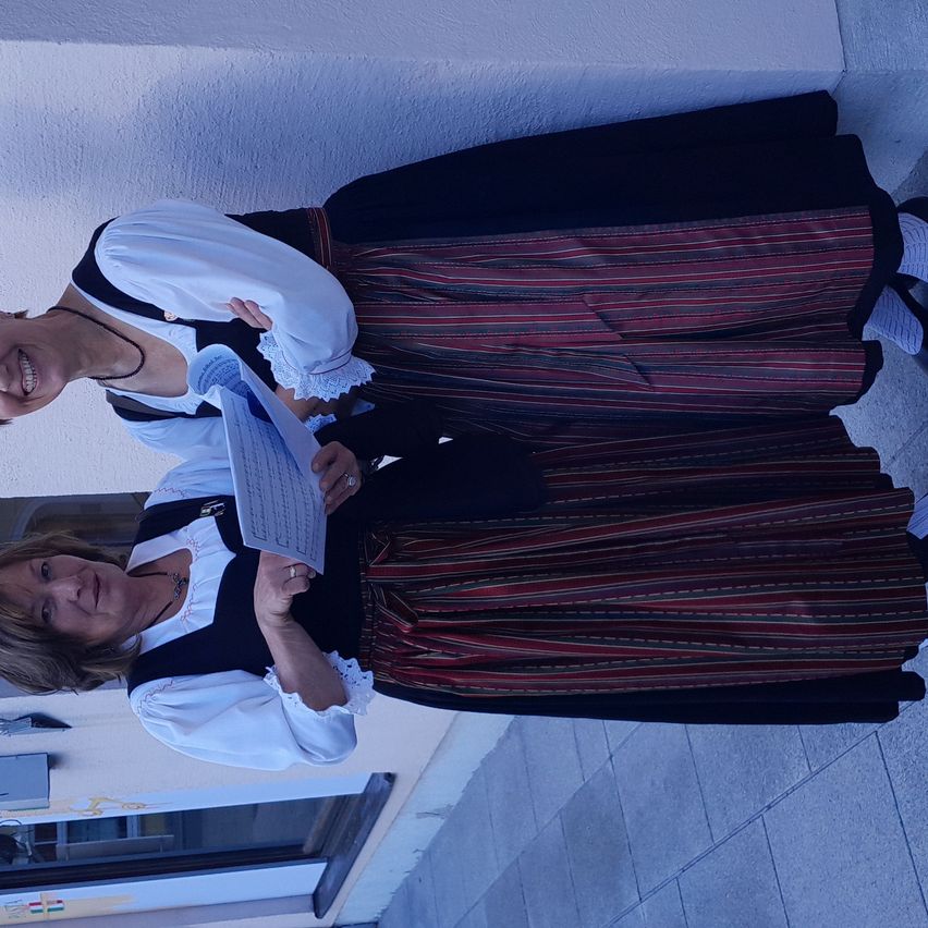 Zwei Frauen in traditionellen Kostümen lächeln, eine hält ein Papier, sie stehen vor einer Wand. Sie tragen Schürzen und Röcke. Der Boden ist gefliest.