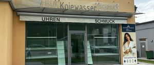 Ein Laden mit Glastüren und Fenstern zeigt ein Schild mit der Aufschrift 'UHREN SCHMUCK'. Neben dem Eingang befindet sich eine Werbung für 'JUBILÄUMSAKTION'. Der Laden ist von einem kleinen Rasenbereich und Gehweg umgeben.