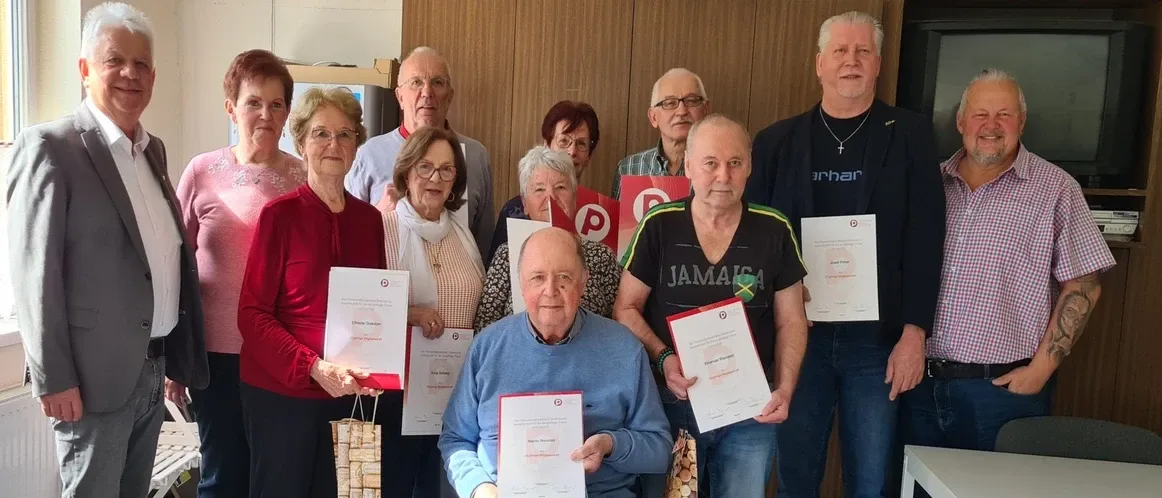 Eine Gruppe älterer Menschen steht und sitzt in einem Raum und hält Urkunden. Ein Mann im Rollstuhl hält eine Urkunde vor sich. Einige Leute tragen Brillen, und ein Mann trägt eine Halskette.