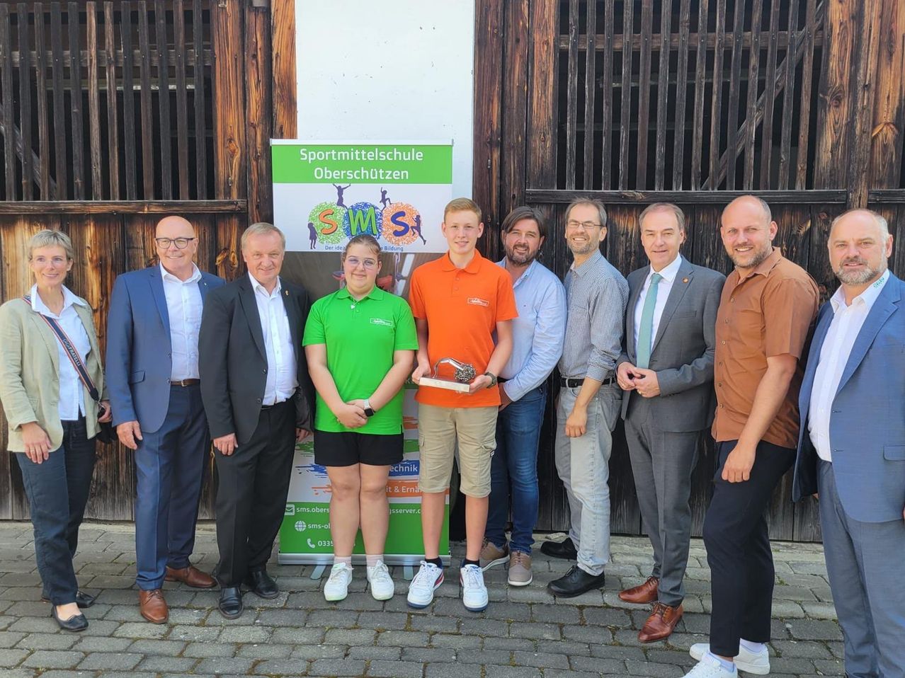 Sieben Personen stehen vor einer Holzstruktur. Ein junges Mädchen in Grün hält einen Pokal. Dahinter hält ein Junge in Orange einen silbernen Pokal. Alle lächeln.