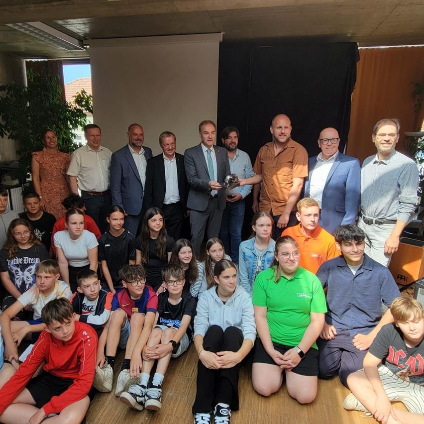 Eine Gruppe von Menschen in formeller Kleidung posiert für ein Foto in einem Raum. Sie stehen und sitzen auf dem Boden. Einige von ihnen halten einen Pokal.