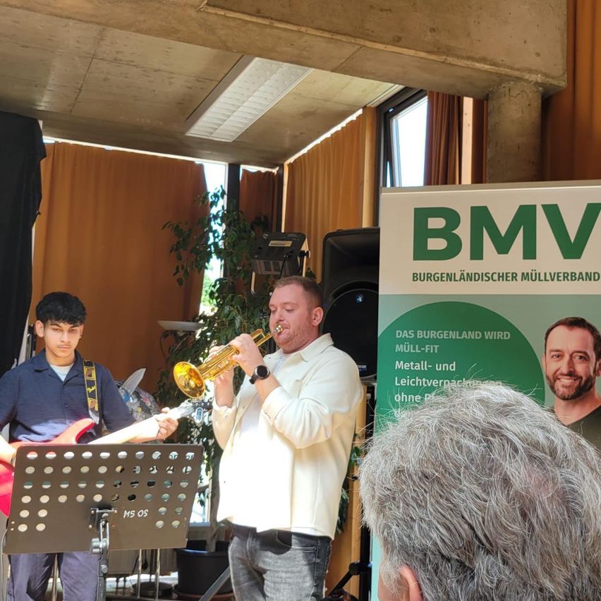 Zwei Musiker spielen auf der Bühne. Ein Mann spielt Trompete, während ein Junge Gitarre spielt. Ein Banner mit dem Text 'BMW' ist im Hintergrund.
