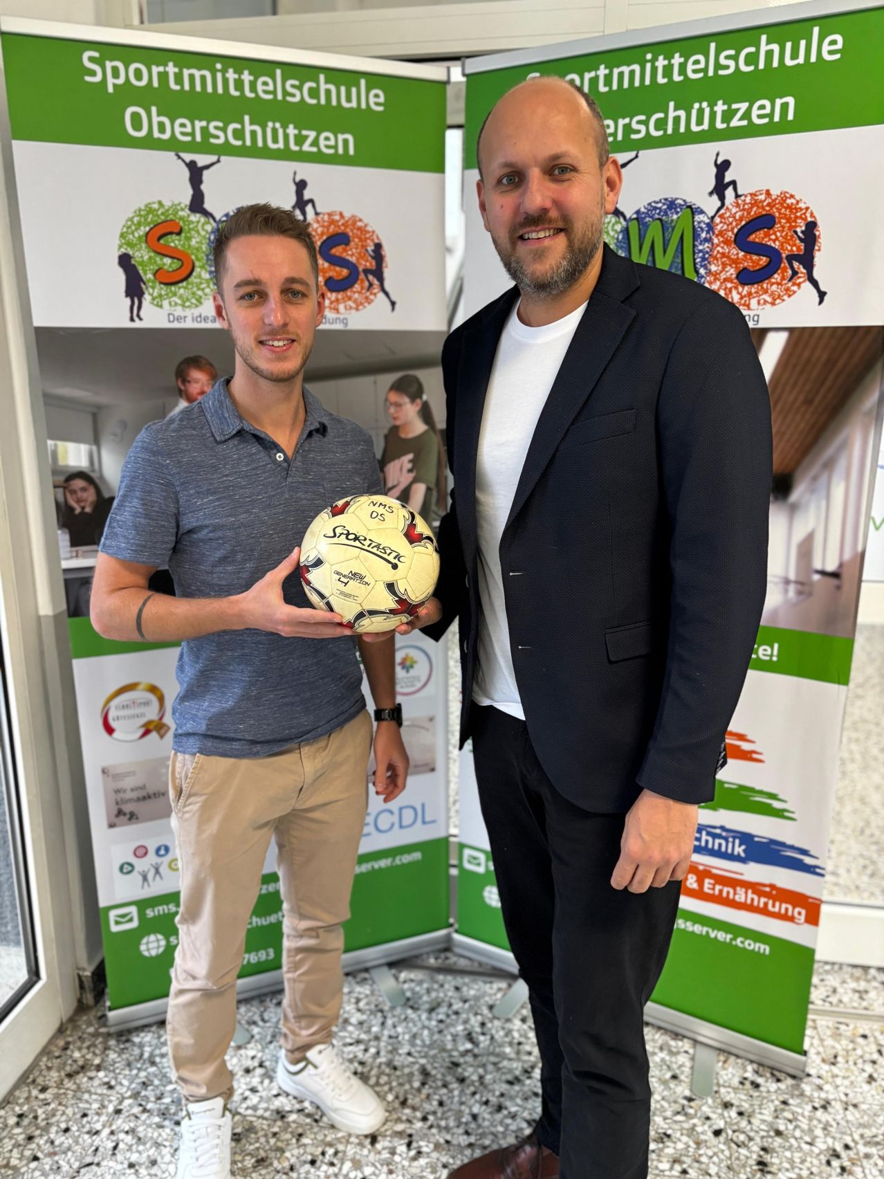 Zwei Männer stehen vor einem Hintergrund mit Sportlogos. Einer hält einen Fußball. Der Mann auf der linken Seite trägt ein blaues Hemd und Khakihosen. Der Mann auf der rechten Seite trägt einen schwarzen Anzug und ein weißes Hemd.