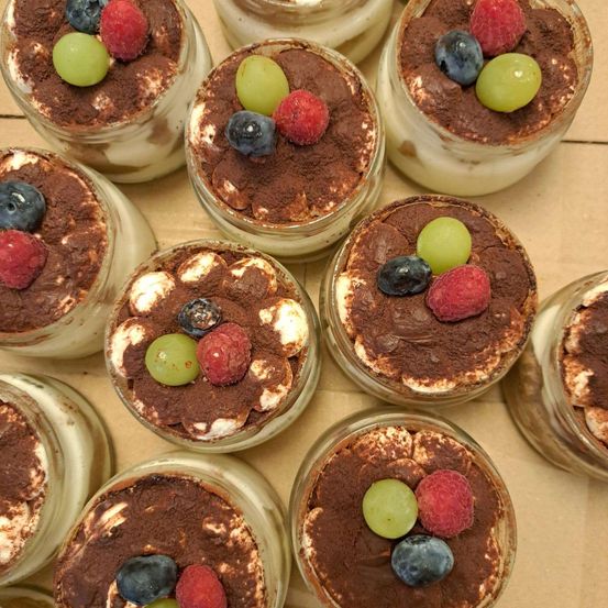 Mehrere Gläser Tiramisu mit Beeren und Trauben obenauf, auf einer Holzoberfläche angeordnet.