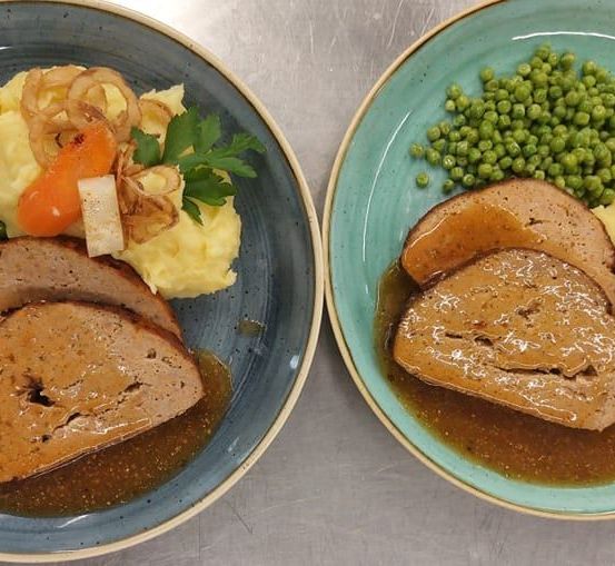Zwei Teller mit Fleisch und Gemüse auf einem Tisch. Beide haben Soße, Erbsen und Karotten. Der linke Teller hat Kartoffelpüree und Zwiebeln.