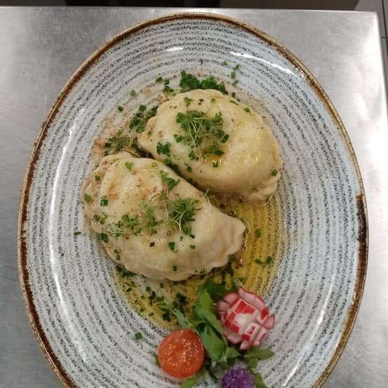 Zwei Knödel auf einem Teller mit Kräutern, einer Tomate und einer Blume. Der Teller steht auf einer metallischen Oberfläche.