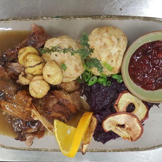 Ein weißer Teller mit Fleisch, Gemüse und Brötchen. Das Fleisch hat Sauce. Daneben sind rote Sauce, Zitrone und eine Beilage.