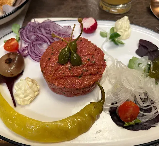 Ein Teller mit einem Fleischpattie, grünem Pfeffer, Kapern, eingelegtem Gemüse und verschiedenen Garnituren auf einem Holztisch.