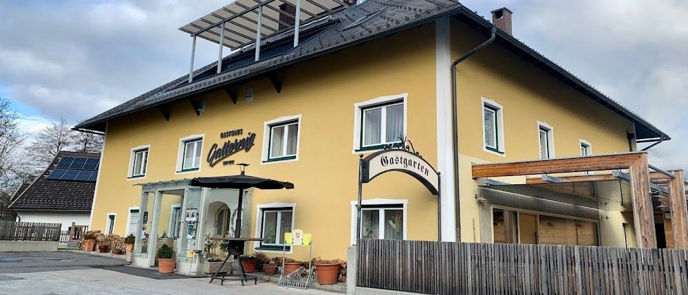 Ein gelbes Gebäude mit mehreren Fenstern und einem Schild mit der Aufschrift Gasthof Gastgarten. Solarmodule sind auf dem Dach, und vor dem Eingang steht ein Regenschirm.