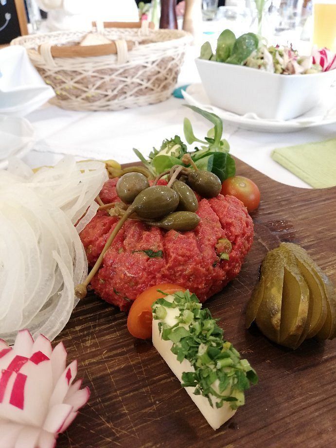 Ein Holzbrett zeigt eine rohe Rinderpastete, die mit Oliven, Gurken und Kräutern belegt ist, mit einem Salatschüssel, einem Korb und einem grünen Tuch im Hintergrund.