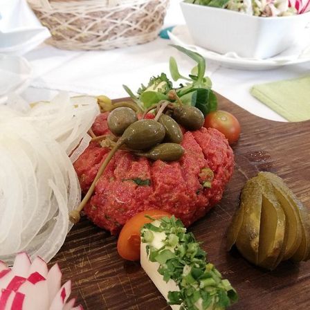 Ein Holzbrett zeigt eine rohe Rinderpastete, die mit Oliven, Gurken und Kräutern belegt ist, mit einem Salatschüssel, einem Korb und einem grünen Tuch im Hintergrund.