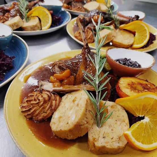 Ein gelber Teller mit Fleisch, Kartoffeln und Gemüse. Das Gericht ist mit Rosmarin und Orangenscheiben garniert. Andere Teller sind ebenfalls auf dem Tisch arrangiert.