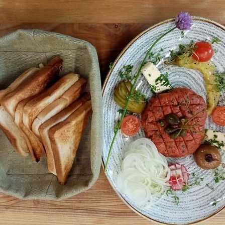 Auf einem Holztisch steht ein Teller mit Fleischpatty, Gurken, Zwiebeln und Tomaten sowie ein Korb mit getoastetem Brot.