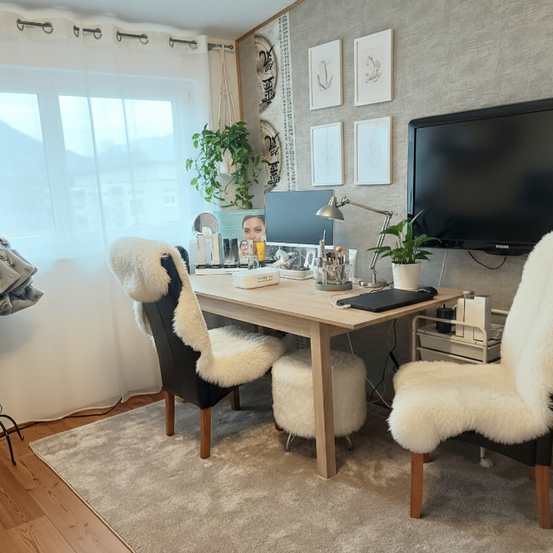 Ein gut beleuchteter Raum mit einem Schreibtisch, zwei Stühlen, einem Monitor und einem Fernseher an der Wand. Der Schreibtisch ist mit einer Lampe, einer Pflanze und verschiedenen Gegenständen geschmückt.