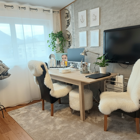 Ein Raum mit einem Schreibtisch, Stühlen und einem Fernseher. Der Schreibtisch hat einen Monitor, eine Lampe, eine Topfpflanze und einige Gegenstände. Es gibt einen Teppich, ein Fenster und gerahmte Bilder an der Wand.