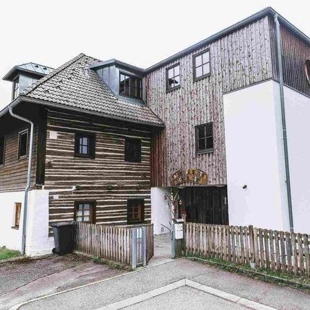 Ein Holzhaus mit mehreren Fenstern und einem kleinen Tor, umgeben von einem Holzzaun und einem Mülleimer.