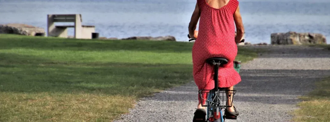 Eine Frau in einem roten Kleid fährt auf einem Kiesweg in der Nähe eines Gewässers mit einer Bank und einem Rasenbereich im Hintergrund mit dem Fahrrad.