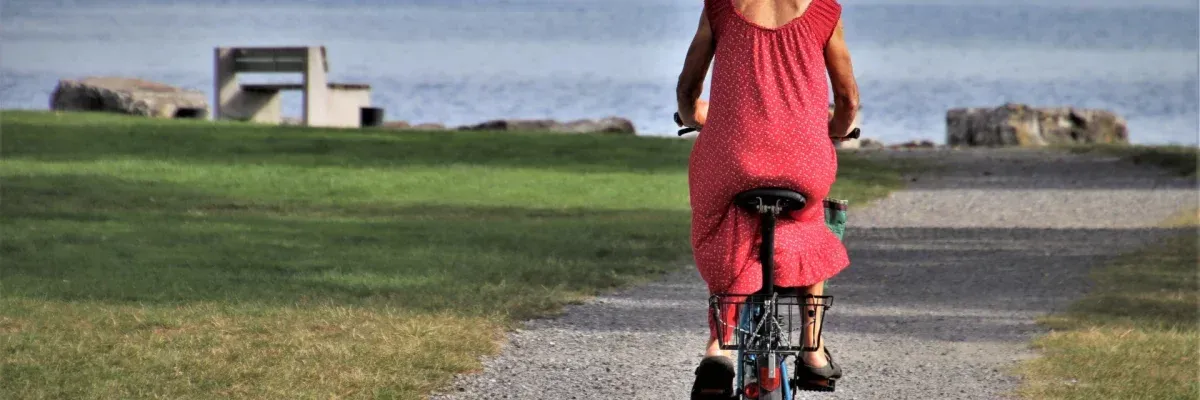 Eine Frau in einem roten Kleid fährt auf einem Kiesweg in der Nähe eines Gewässers mit einer Bank und einem Rasenbereich im Hintergrund mit dem Fahrrad.