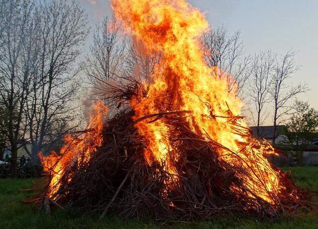 Ein Lagerfeuer brennt hell in einem Feld mit einigen kahlen Bäumen und einem Haus im Hintergrund.