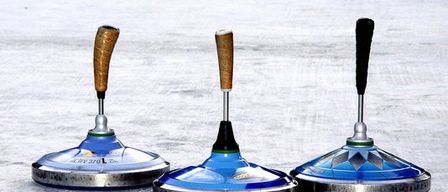 Drei blaue Curlingbesen mit braunen Griffen liegen auf einer verschneiten Eisbahn, mit einem schwarzen Puck in der Ferne.