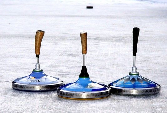 Drei blaue Curlingbesen mit Holzgriffen stehen auf einer vereisten Oberfläche. Die Besen haben silberne Basen und schwarze Griffe. Im Hintergrund befindet sich ein schwarzer Puck auf dem Eis.