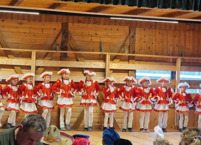 Eine Gruppe junger Mädchen in passenden roten und weißen Kostümen tritt auf der Bühne auf. Sie stehen in einer Reihe und schauen das Publikum an. Die Bühne ist aus Holz, und es gibt Vorhänge im Hintergrund. Ein Publikum beobachtet sie.