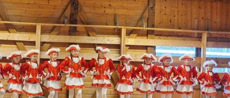 Eine Gruppe junger Mädchen in passenden roten und weißen Kostümen tritt auf der Bühne auf. Sie stehen in einer Reihe und schauen das Publikum an. Die Bühne ist aus Holz, und es gibt Vorhänge im Hintergrund. Ein Publikum beobachtet sie.