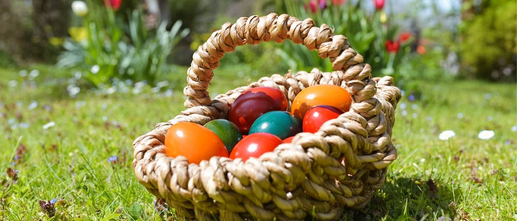 Ein geflochtener Korb mit bunten Ostereiern auf einem Rasen mit Blumen im Hintergrund.
