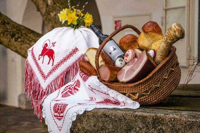 Ein Korb aus Weide gefüllt mit Brot, Fleisch und einer Flasche Wein, geschmückt mit einem rot-weißen Tuch, steht auf einer Steinfläche mit gelben Blumen darüber.