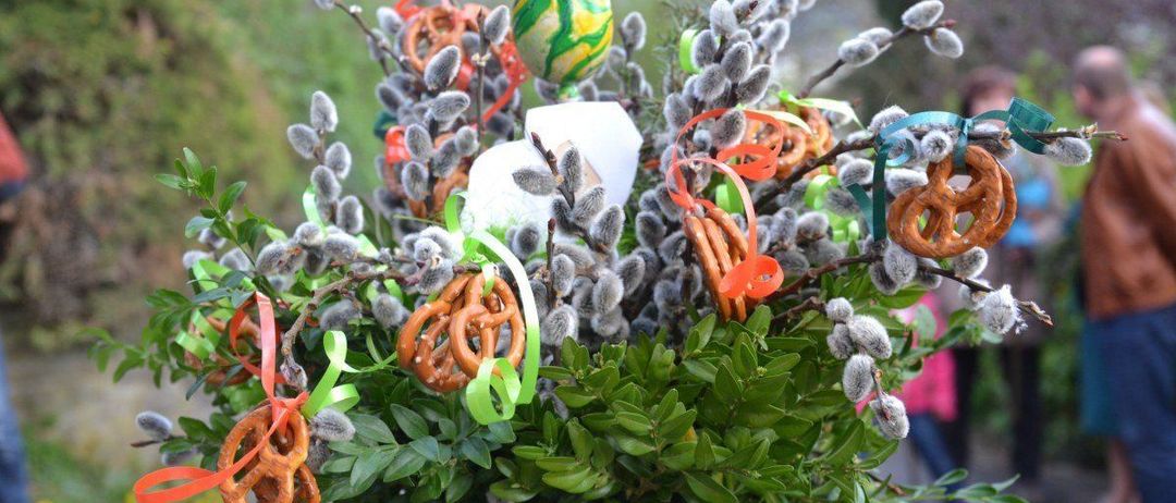 Eine Blumenarrangement mit Ostereiern, Brezeln und bunten Bändern. Das Arrangement enthält grüne Blätter und weiße Weidenkätzchen.