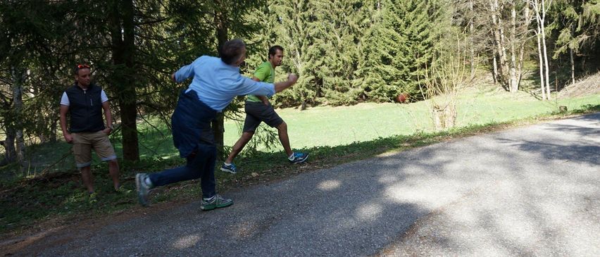 Zwei Männer joggen auf einem Kiesweg, einer in einem blauen Hemd und der andere in einem grünen Hemd. Sie sind von Bäumen und Gras umgeben.