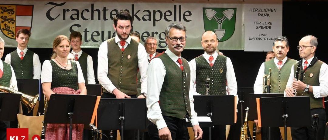 Eine Gruppe von Musikern in traditioneller Kleidung steht auf der Bühne, verschiedene Instrumente sind sichtbar, und ein Banner im Hintergrund.