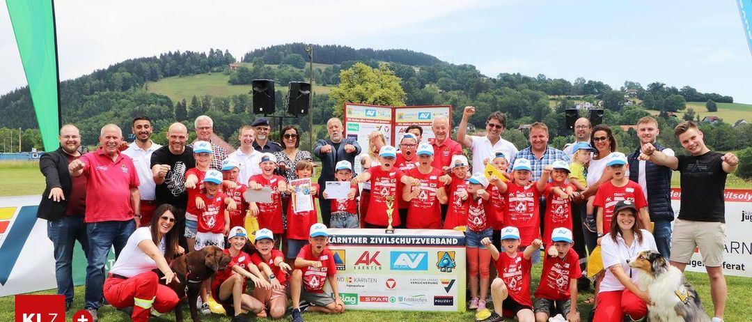Eine Gruppe von Menschen, einschließlich Kindern und Erwachsenen, posiert für ein Foto auf einem Rasenfeld. Sie tragen rote T-Shirts mit Logos. Einige halten Urkunden und Pokale. Hinter ihnen steht ein Banner mit Logos und Text. In der Ferne gibt es Bäume und einen Hügel.