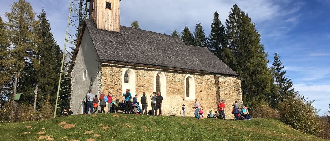 Eine kleine Kirche mit steinerner Außenwand und Spitzdach steht auf einem grasbewachsenen Hügel, umringt von Menschen. Hohe Bäume umgeben die Kirche, und ein Kommunikationsturm steht in der Nähe.
