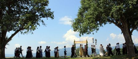 Eine Gruppe von Menschen in traditioneller Kleidung geht in einer Reihe unter einem Baldachin auf einem Rasenfeld. Einige tragen Laternen. Bäume und Berge sind im Hintergrund.
