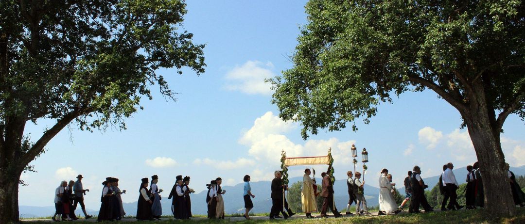 Eine Gruppe von Menschen in traditioneller Kleidung geht in einer Reihe unter einem Baldachin auf einem Rasenfeld. Einige tragen Laternen. Bäume und Berge sind im Hintergrund.