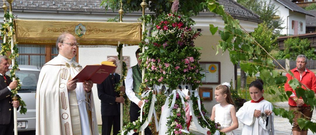 Ein Priester führt eine Zeremonie auf einem Kirchhof durch. Er liest aus einem Buch, während ein mit Blumen geschmückter Baum hinter ihm steht. Zwei junge Mädchen halten Bänder. Dahinter ist ein Gebäude und Bäume zu sehen.