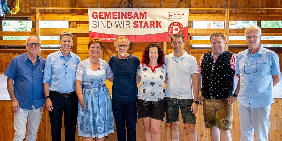 Eine Gruppe Erwachsener posiert für ein Foto vor einer Holzwand mit einem Banner, auf dem 'GEMEINSAM SIND WIR STARK' steht.