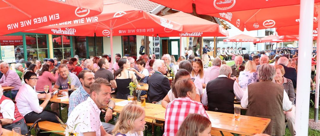 Eine Gruppe von Menschen, darunter Männer, Frauen und Kinder, sitzt unter roten Sonnenschirmen an Tischen in einem Restaurant im Freien. Einige Leute trinken Bier und andere unterhalten sich. In der Ferne stehen einige Personen in der Nähe eines Gebäudes.